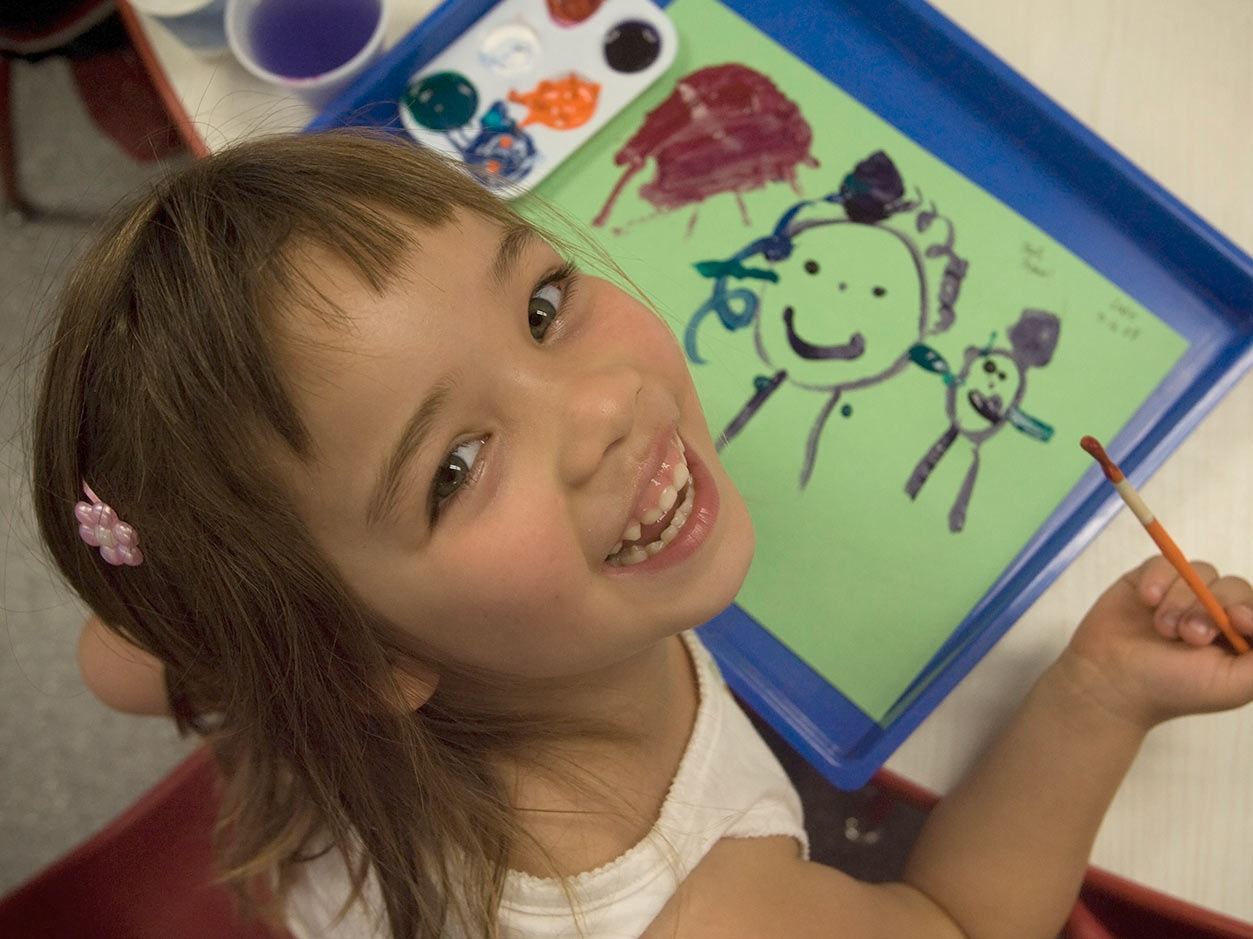an image of a girl smiling up at the camera with a paint bush in her hand and a painting beneath her hand on the table